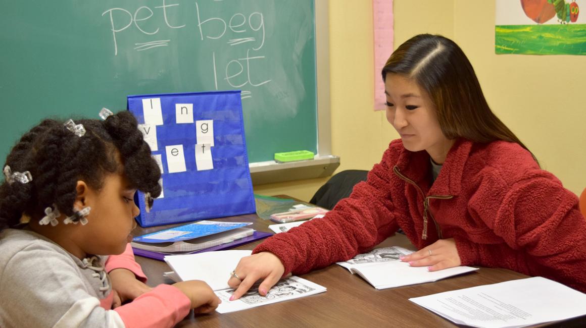 Duquesne student helping young learner read book at table with coordinated visual on table