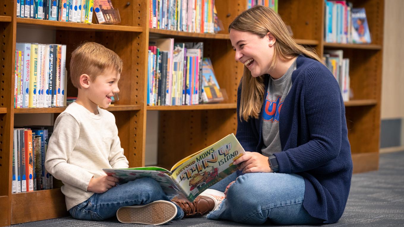 Duquesne Reading Specialist Certificate student and young learning smiling at each other while reading and sharing book on library floor to work on literacy education