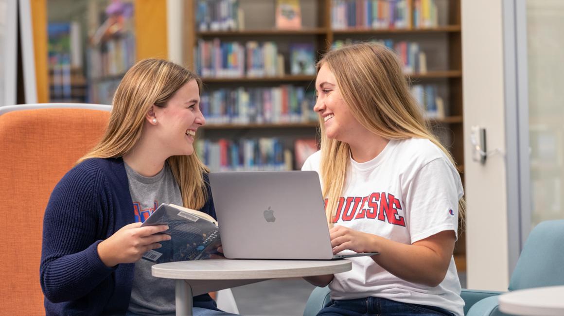 Two Duquesne students conversing while in library, one has a book and the other has a laptop