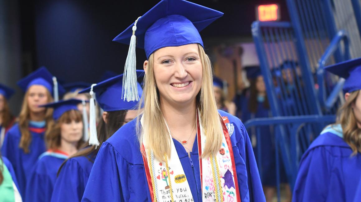 Scholar graduate at commencement in gradaution regalia smiling in cap and gown with color graduate regalia around neck with yound student drawings and notes in colorful marker.