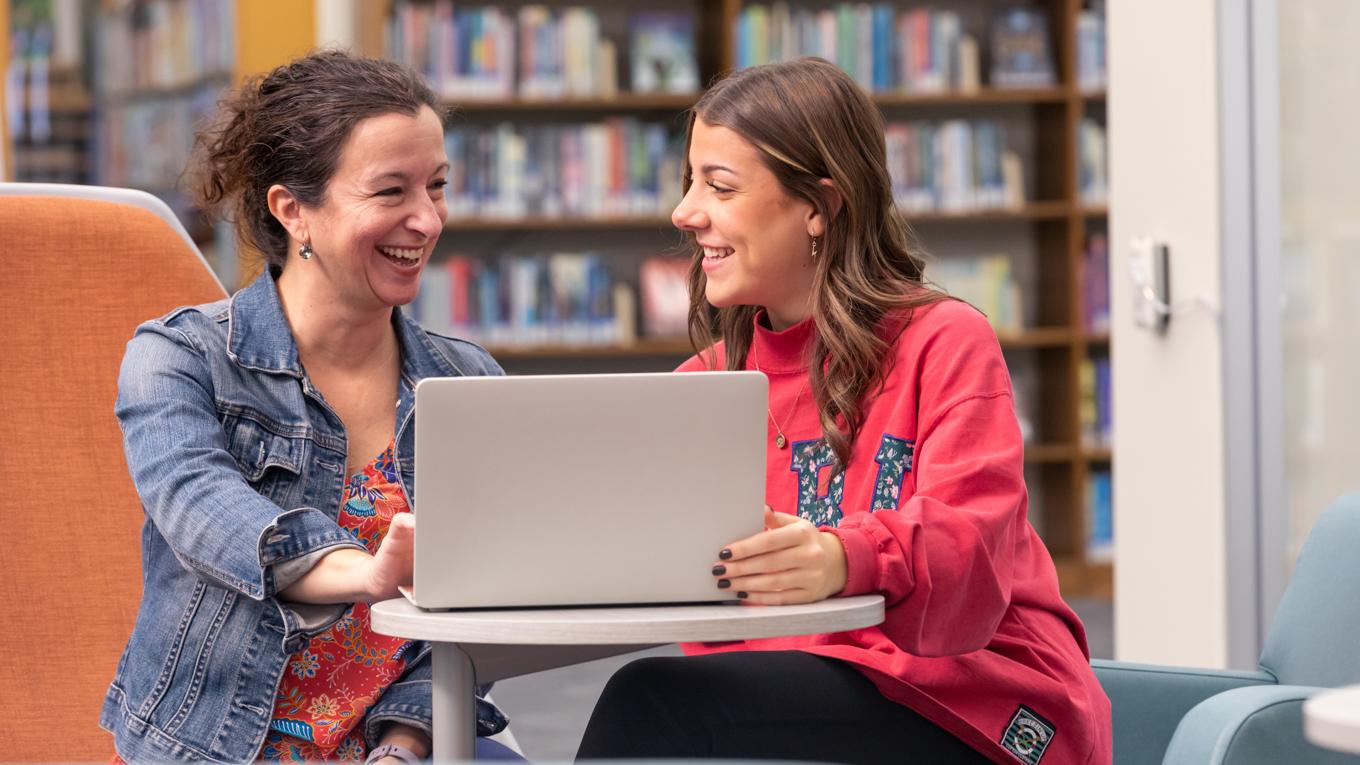 Student and teacher working togehter, smiling, and conversing around laptop in library