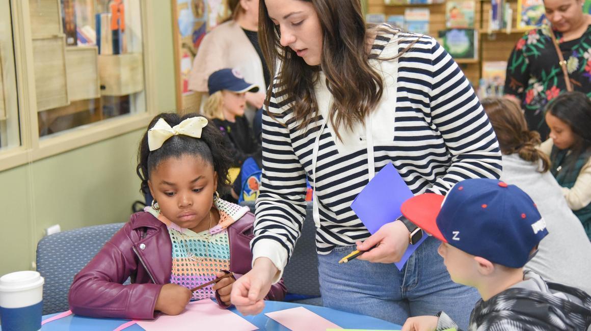 School of Education student teacher in field in library engaging elementary students in hands-on experience in construction paper lesson.