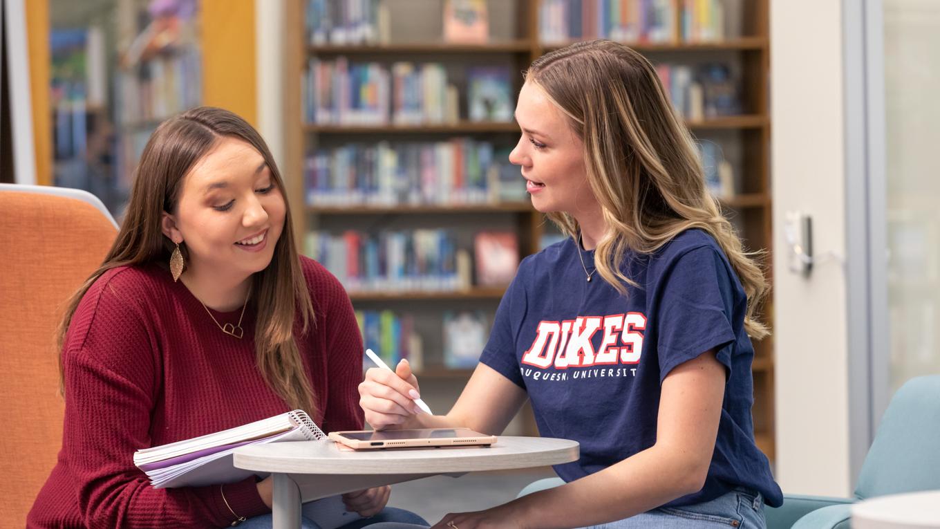 Two Duquesne MAT Secondary Education 7-12 students working on classroom curriculum in library with paper and electronic tablet and bookshelves in background