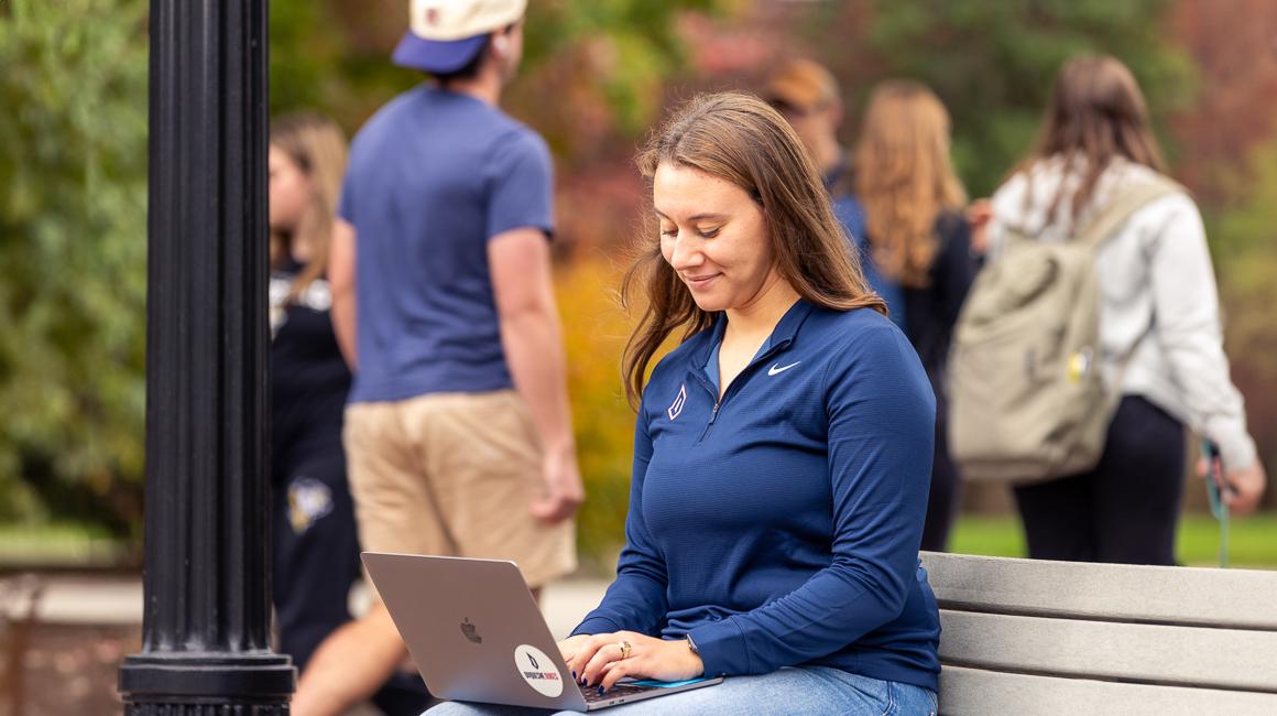 Secondary Education MAT Duquesne studing taking online classes on laptop sitting on bench outside of campus with campus activity in background