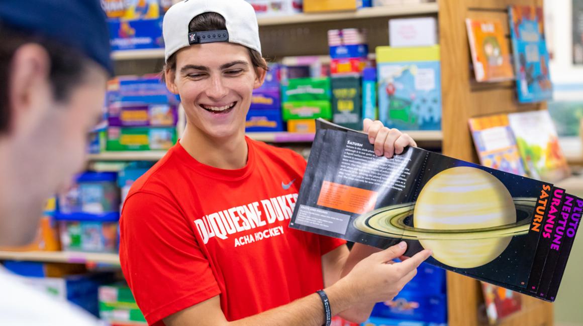 Student smiling and reading children's science picture book with colleagues in Curriclum Center library