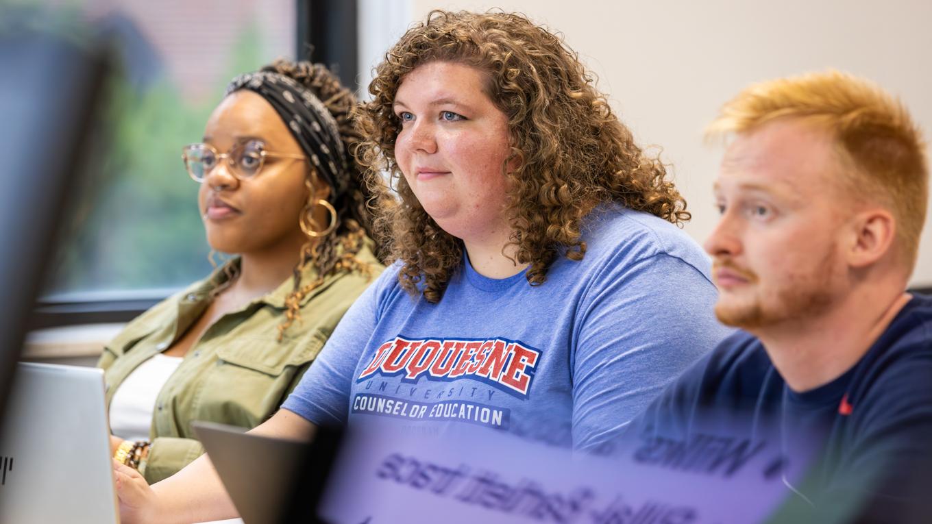 Three Ph.D. Counselor Education and Supervision students engaged listening to classroo discussion at desk with laptops and laptop reflection artistically displayed in location of desk