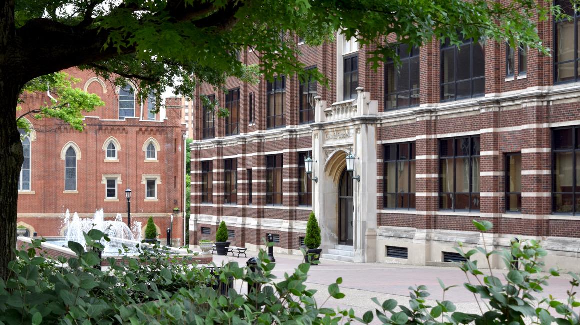  Tree-framed Canevin Hall and to the side the Administration building with the water fountain in the center of the campus walkway and framed by the trees.