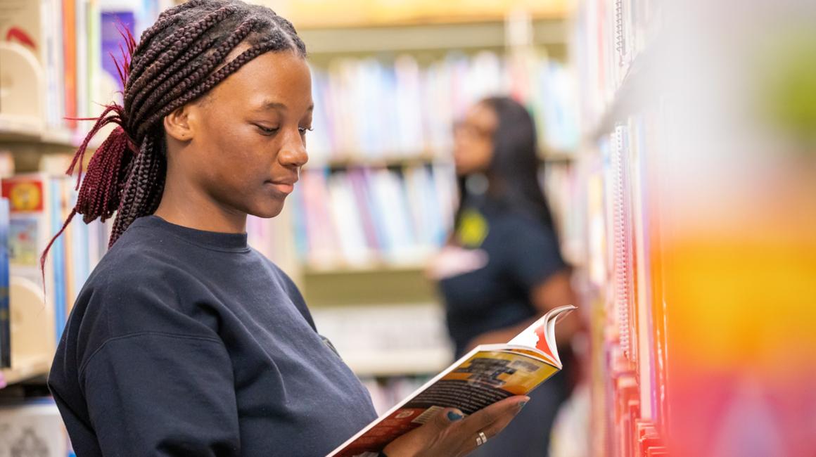 Student reading book from library shelf and other student walking behind her in background