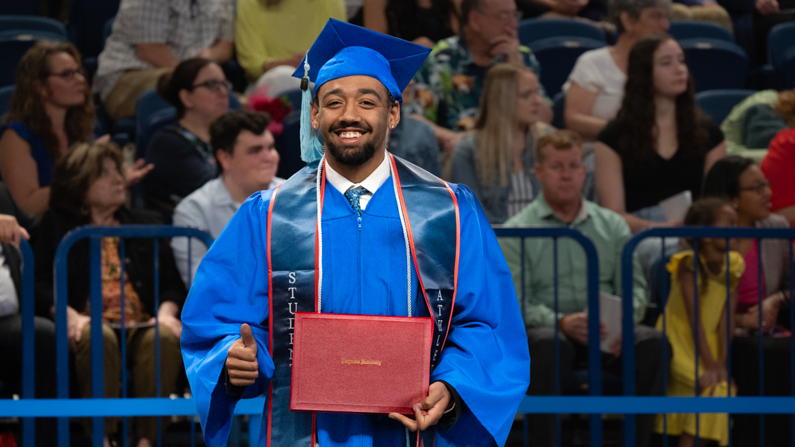 School of Education B.S.Ed. Secondary Education alum Ezekiel Daure smiles and gives a thumbs-up with diploma in-hand and commencement crowd behind him