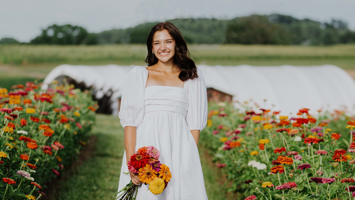 School of Education student Italia Cicione holding flowers with a field of flowers around her and a greenhouse and mountain behind her