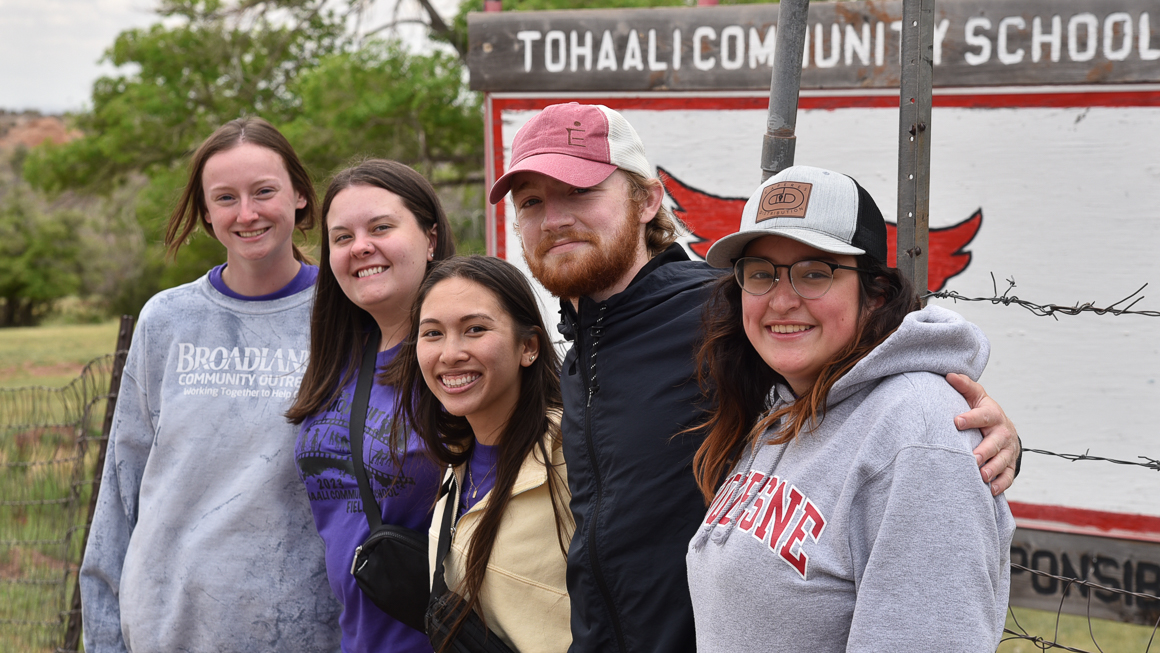 Photo of five School of Education students in New Mexico smiling in group outside with Tohaali Community School sign behind them