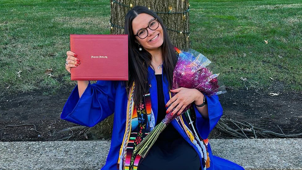 Sarah Lopez sitting on campus wearing graduation regalia holding diploma and flowers in hands smiling