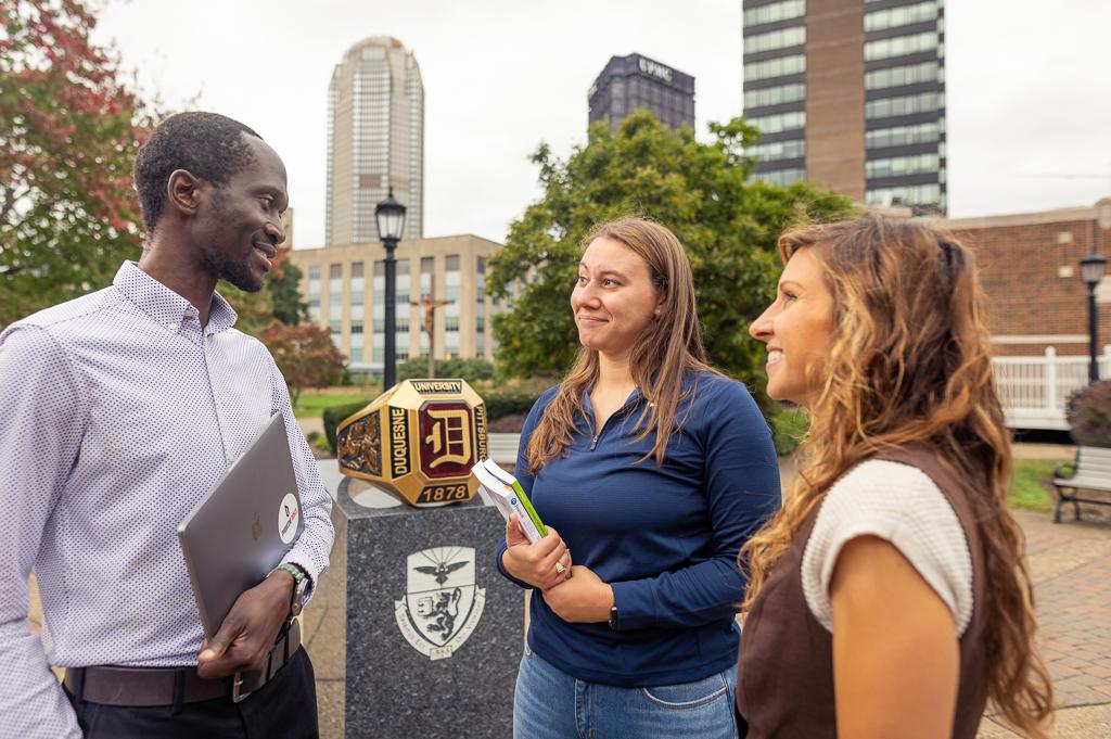 Three graduate students conversing on campus with books and laptop and ring monument in background students walkng into campus building