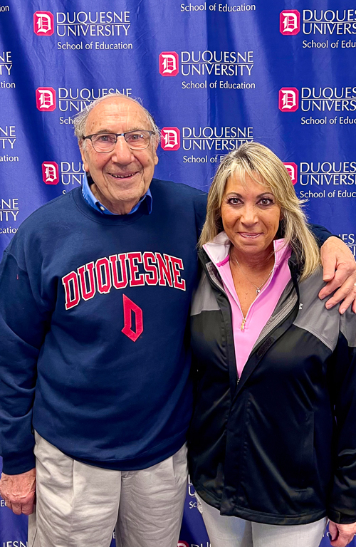 Photo of alum Alex Joseph wearing a Duquesne sweater smiling with his arm around his daughter next to him and a Duquesne backdrop behind him