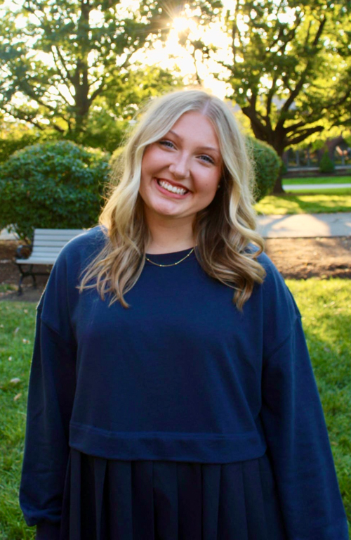 Elle Bowers B.S.Ed. Dual Certification in Early Childhood Education & Special Education PreK-12 student smiling with sunlit trees and bench outside on campus in background.