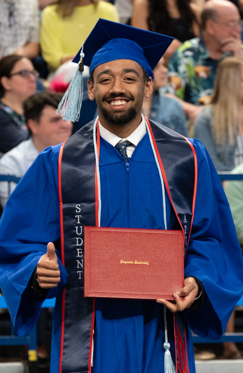 School of Education B.S.Ed. Secondary Education alum Ezekiel Daure smiles and gives a thumbs-up with diploma in-hand and commencement crowd behind him