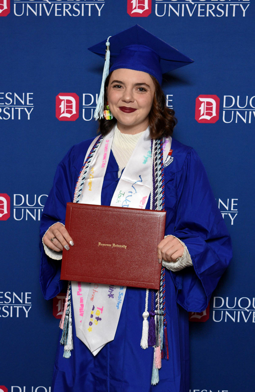 Grace Barcaskey holding diploma and smiling in graduation regalia during commencement with Duquesne University backdrop print in background.