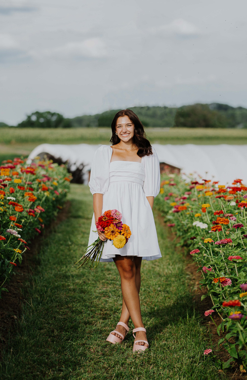 School of Education student Italia Cicione holding flowers with a field of flowers around her and a greenhouse and mountain behind her