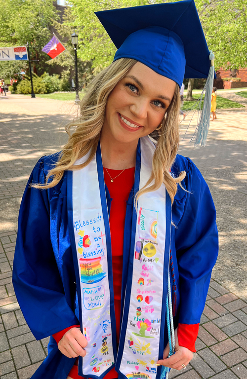 Jayne Simon Aulicino in graduation regalia outside on campus with campus activity in background