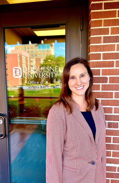 Photo of M.S. Higher Education Administration alum Katy Tersine outside door with Duquesne University Office of Residence Life sticker on it and reflection of campus buildings reflected in door glass