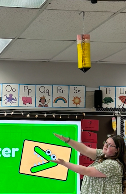 B.S.Ed. Dual Certification in Early Childhood Education & Special Education PreK-12 student Kendall House teaching greater than math lesson in front of screen with blackboard behind in field experience classroom.
