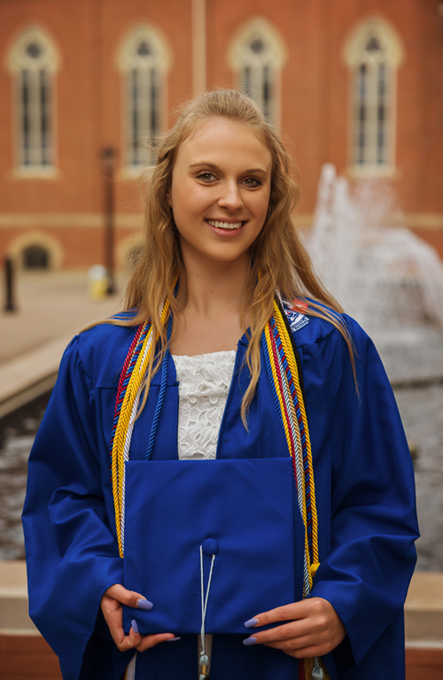 Lauren Barnes in graduation regalia outside on campus with fountain and building behind her