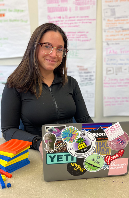 Photo of B.S.Ed. Middle School undergraduate student Lila Scheer smiling at desk with laptop and learning tens blocks in from of her and info posters behind her