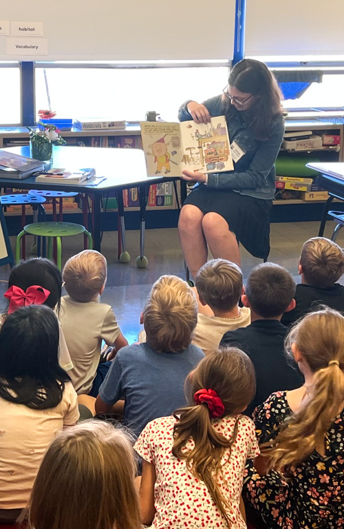 Early childhood education student Maura Riscavage on chair reading to group of classroom students seated on floor during hands-on field experience.