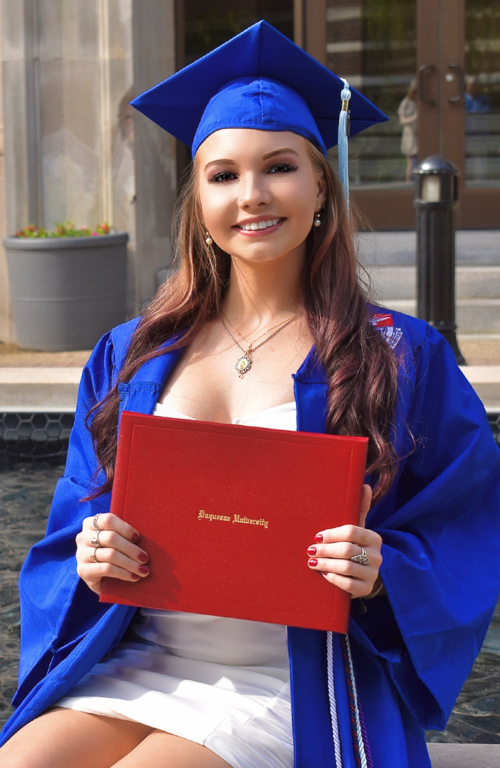 Photo of B.S.Ed. Middle Level Education alum Natasha Cote in graduation regalia smiling as she holds her diploma outside the School of Education's Canevin Hall
