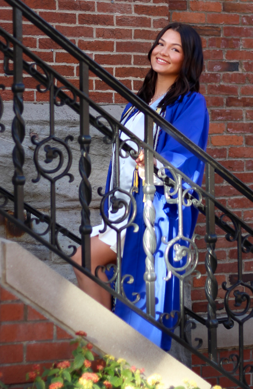 Photo of Rachel McGough on steps of Administration building in graduate regalia holding the railing and smiling