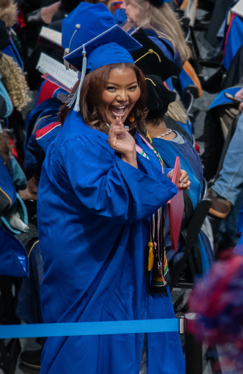 Photo of Venetia on commencement walking floor at with her undergraduate diploma in hand smiling at crowd member who is cheering her on with pom pom and new graduates are seating in the background.