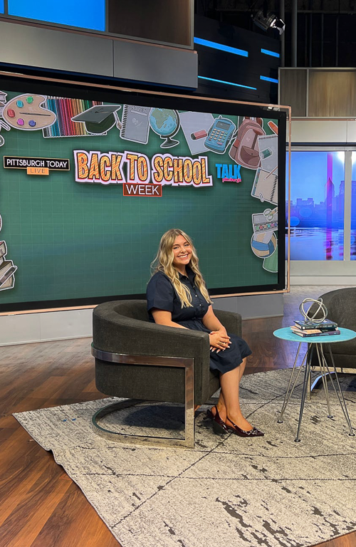School of Education student Zoe Brunick on KDKA Talk Pittsburgh set for Teach Me Tuesday show seating in chair with table with books in front and backdrop of TV show titles reading Pittsburgh Today Live; Back to School Week; Talk Pittsburgh.