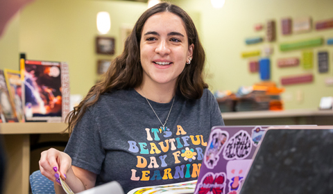 School of Education student wearing It's a Beautiful Day for Learning t-shirt as she is smiling and turning the page of a book from behind her laptop in the library curriculum center