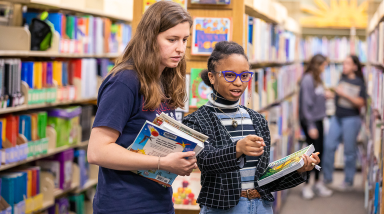 School of Education students exploring resources in the library's curriculum center with books in hand and colorful bookshelves and student activity behind them