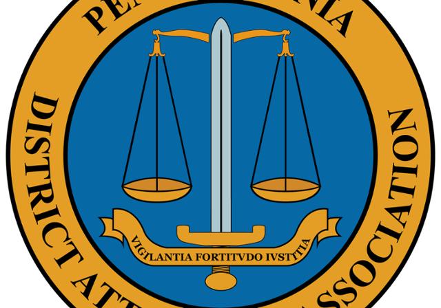 Pennsylvania District Attorneys Association logo with those words written in black text around the outside of a gold circle.  A scales of justice is in the middle of that circle, placed against a medium blue background.