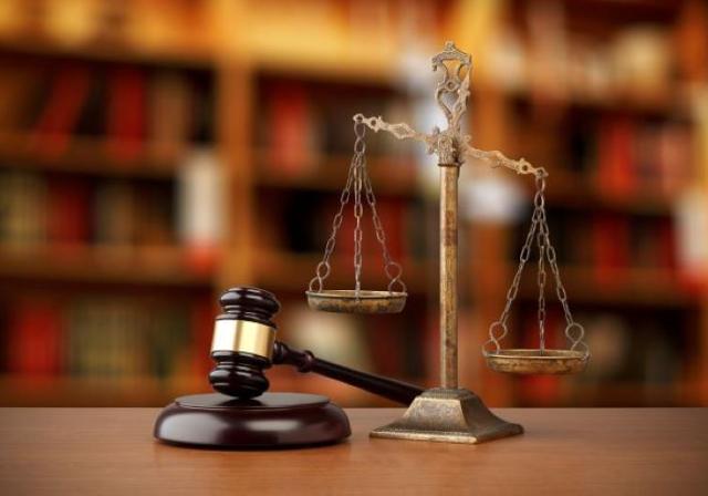 A gold scales of justice and brown wooden gavel on a desktop with faded bookshelves in the background.