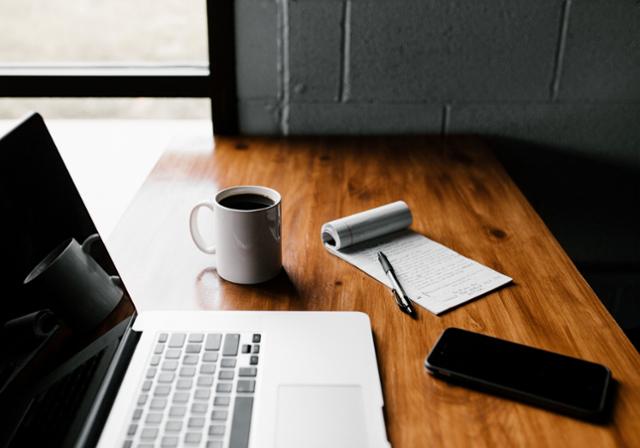 An open laptop, white coffee mug, black cell phone, and small writing tablet on top of a wooden table top.