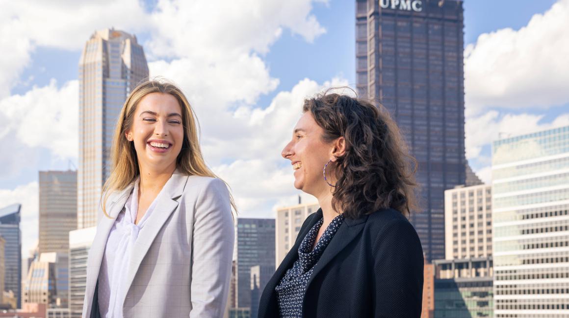 Two students, dressed in business attire, talking outside with the Pittsburgh skylin behind them in the distance.