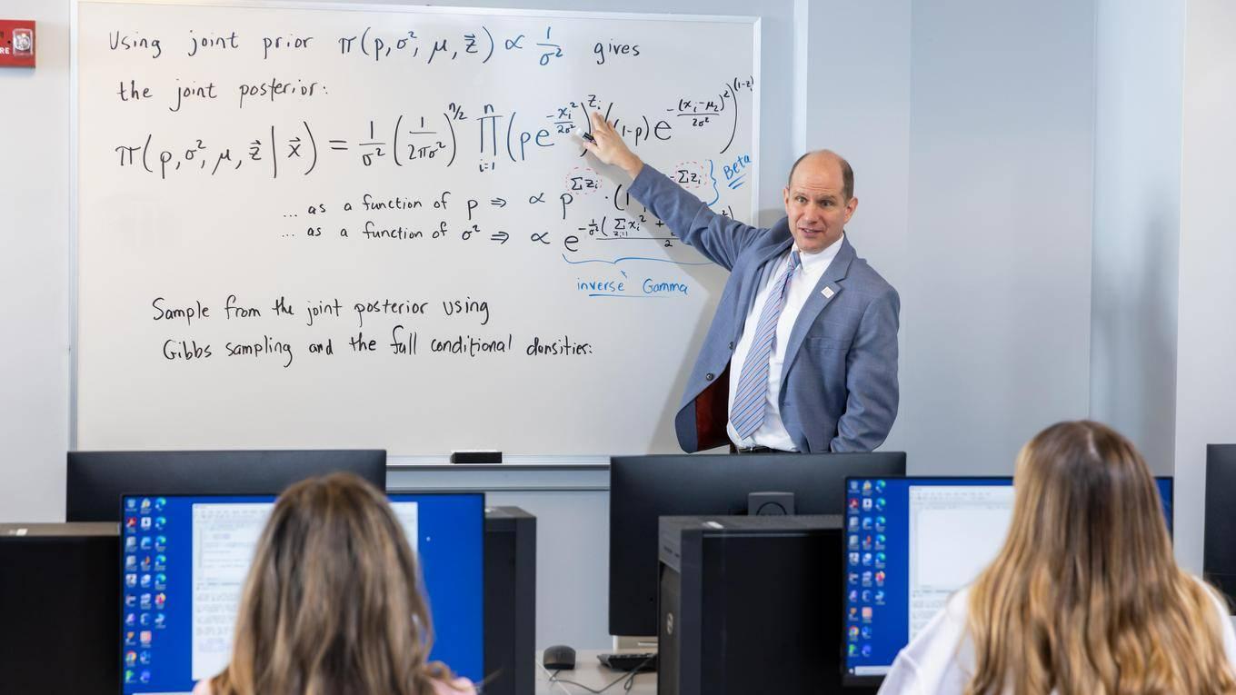 Professor at the white board teaching Gibbs sampling to two students sitting by desktop computers