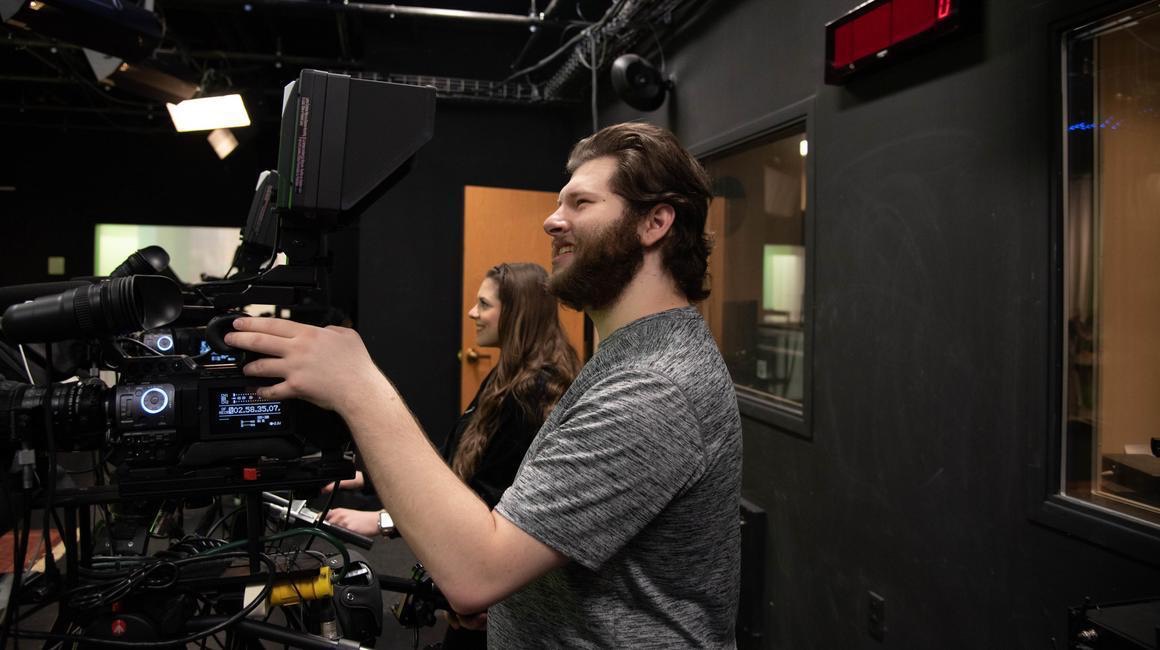 A student operates a professional broadcast camera in College Hall's media studio, smiling as a second student looks on in the background.