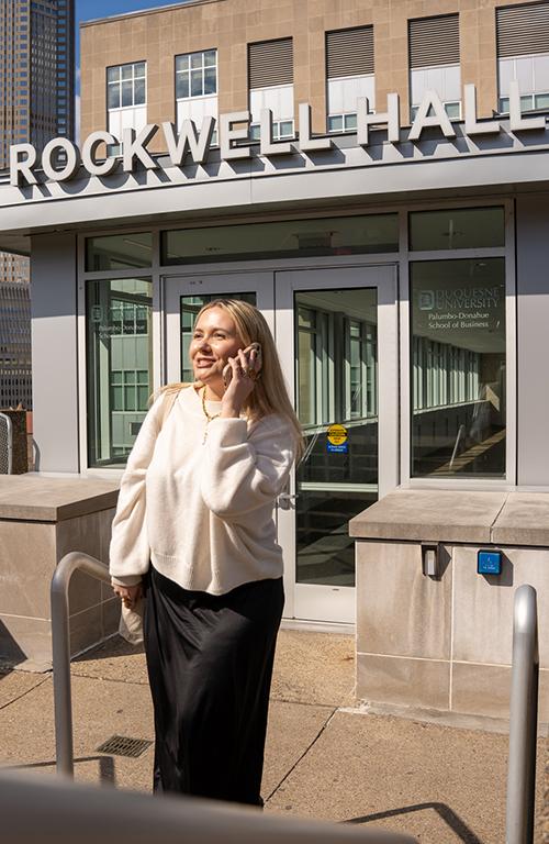 Jayda Benson emerging from Rockwell Hall on her cell phone, sunshine on her face. 