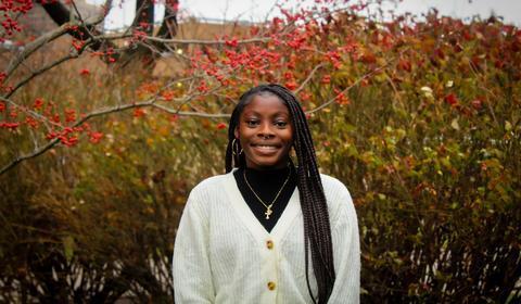 A smiling young woman with braided hair, wearing a cream cardigan and gold jewelry, standing outdoors in front of red berry bushes.