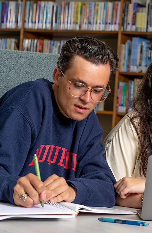 A Duquesne University student wearing a navy blue Duquesne sweatshirt and glasses writes in a spiral notebook while seated next to another student in a library, bookshelves visible in the background.
