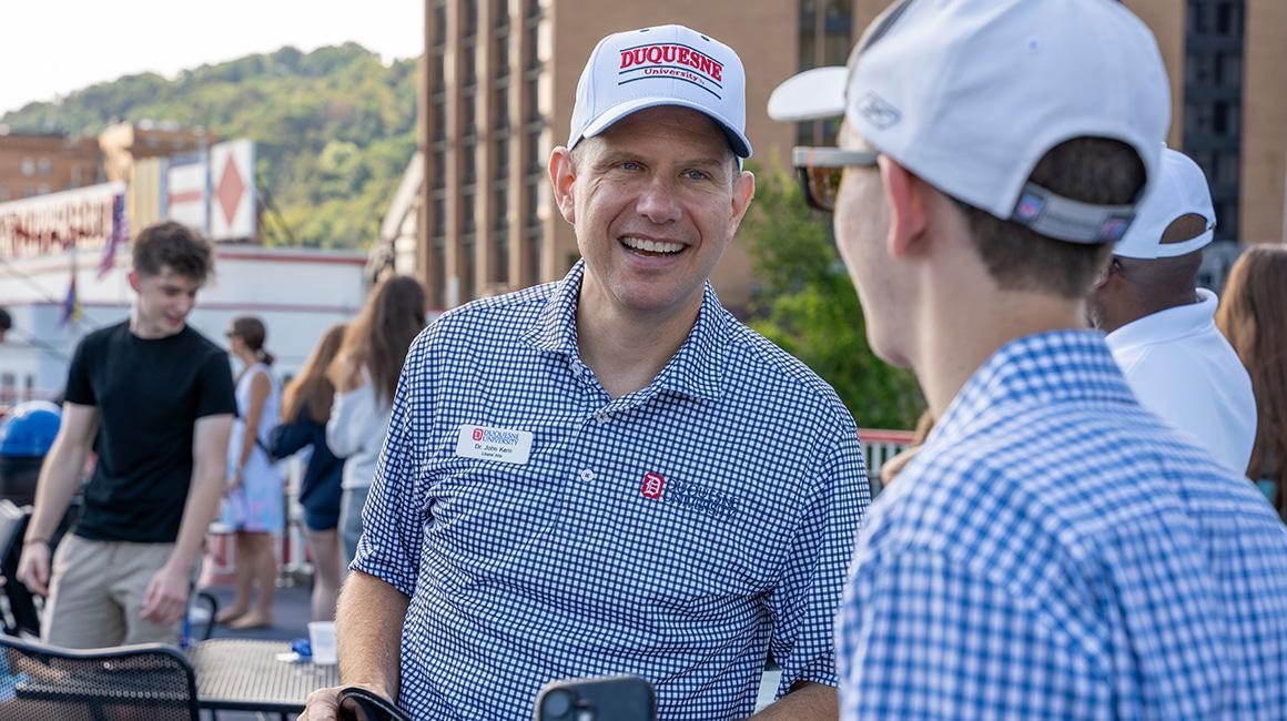Associate dean wearing a Duquesne University cap and checkered shirt smiles while talking with a student on the deck of the Gateway Clipper during orientation, with the Pittsburgh hillside and riverfront buildings in the background.