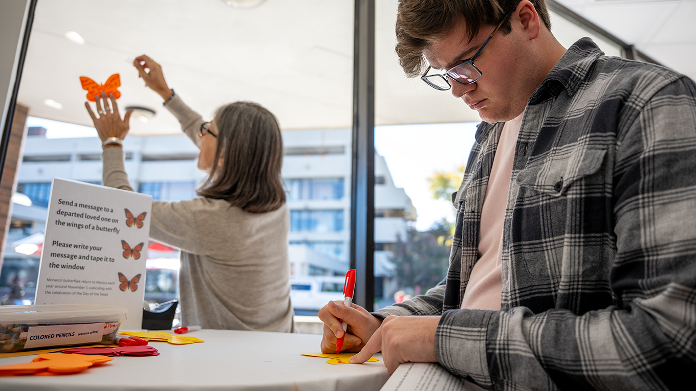 Student writes message to loved one during Día de los Muertos celebration. Professor hangs messages on window.