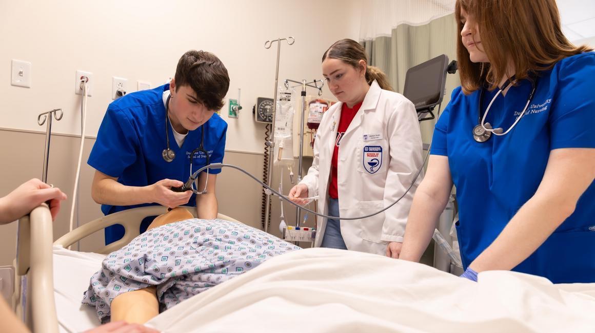 Three students surround a mannequin in a hospital bed.