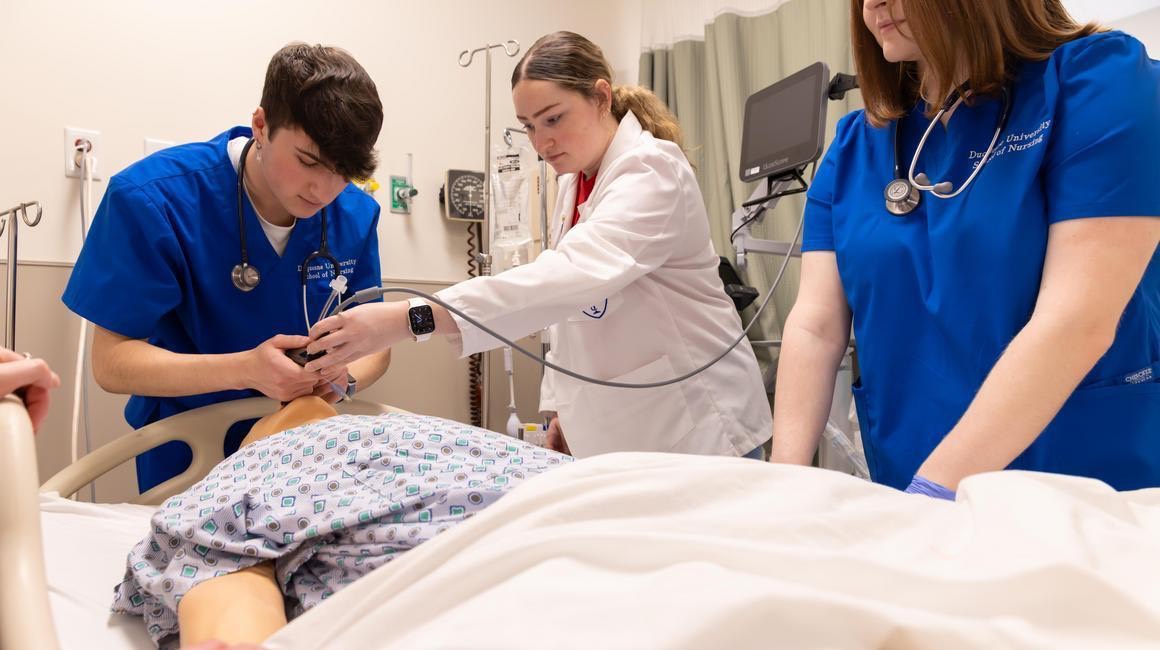 Three students in scrubs practice clinical skills on a medical simulation mannequin in a nursing lab.