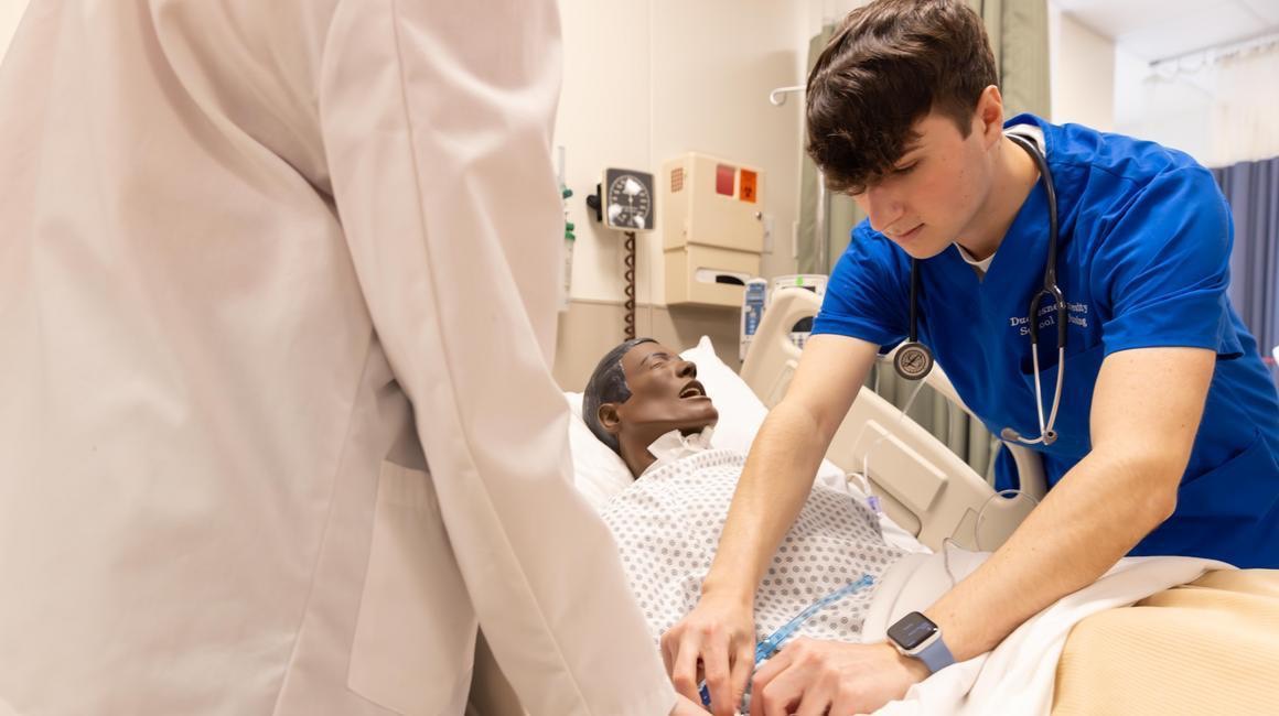 Student in scrubs and student in white coat practice in nursing lab on training mannequin.