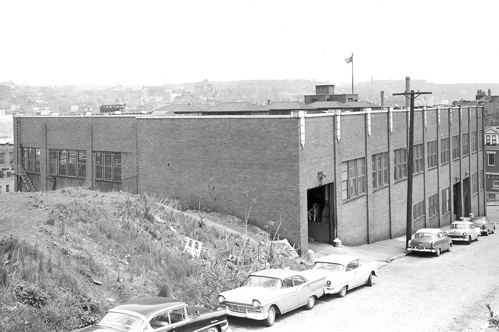 Historic exterior view of the Duquesne University music building prior to renovation, showing a brick industrial structure on the Bluff with parked mid-century cars and the Pittsburgh skyline in the background.