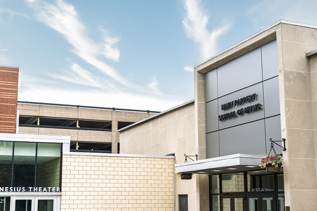 Exterior view of the Mary Pappert School of Music at Duquesne University, showing the main entrance with updated stone façade, signage, and the Genesius Theater entrance adjacent under a bright sky.